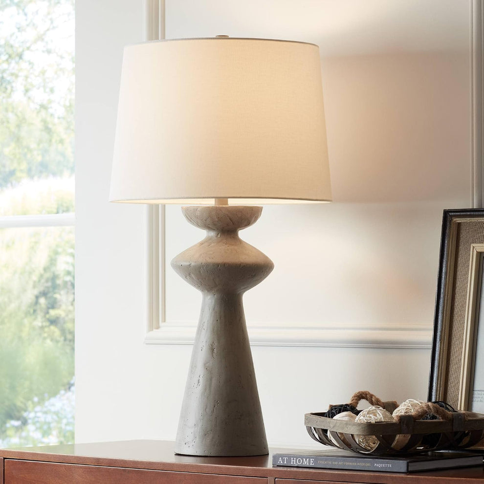 Grey sculptural table lamp with white drum shade on a wooden console in a softly lit room