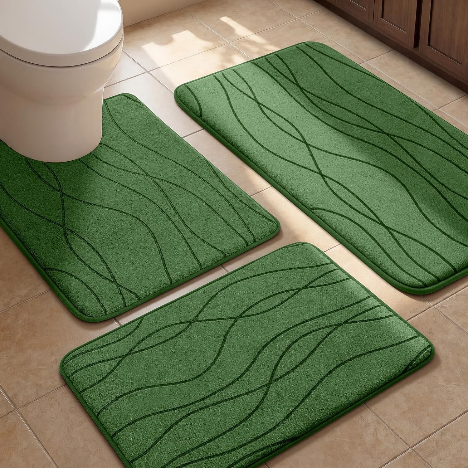 Three green memory-foam bath mats with wavy embossed lines arranged on a tiled bathroom floor near a toilet and cabinet.