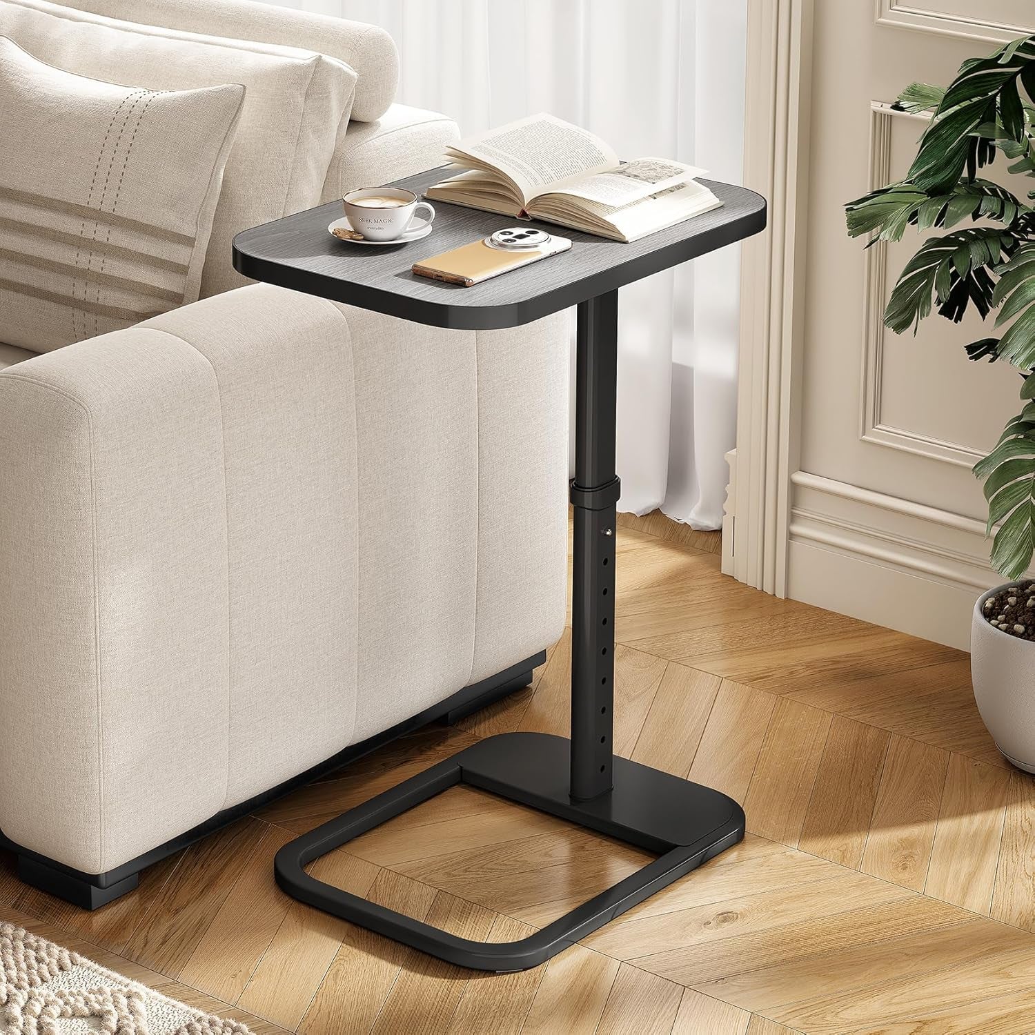 C‑shaped black metal side table beside a beige sofa holding a cup, book and phone