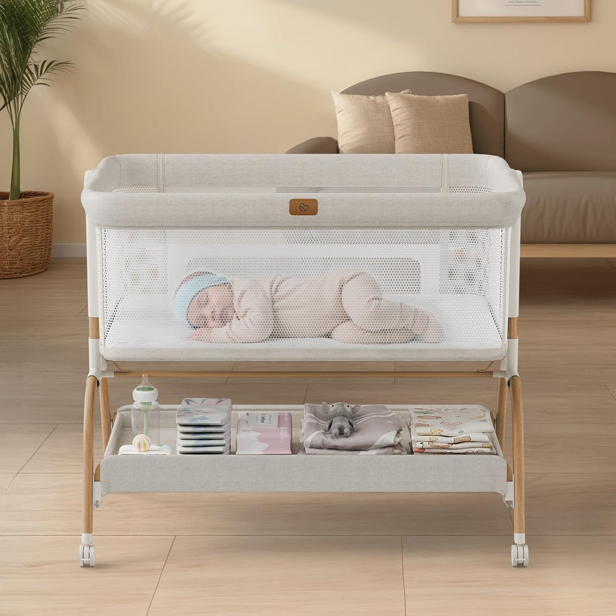 Front view of a beige bedside bassinet with mesh sides, a sleeping baby inside and organized storage shelf below.