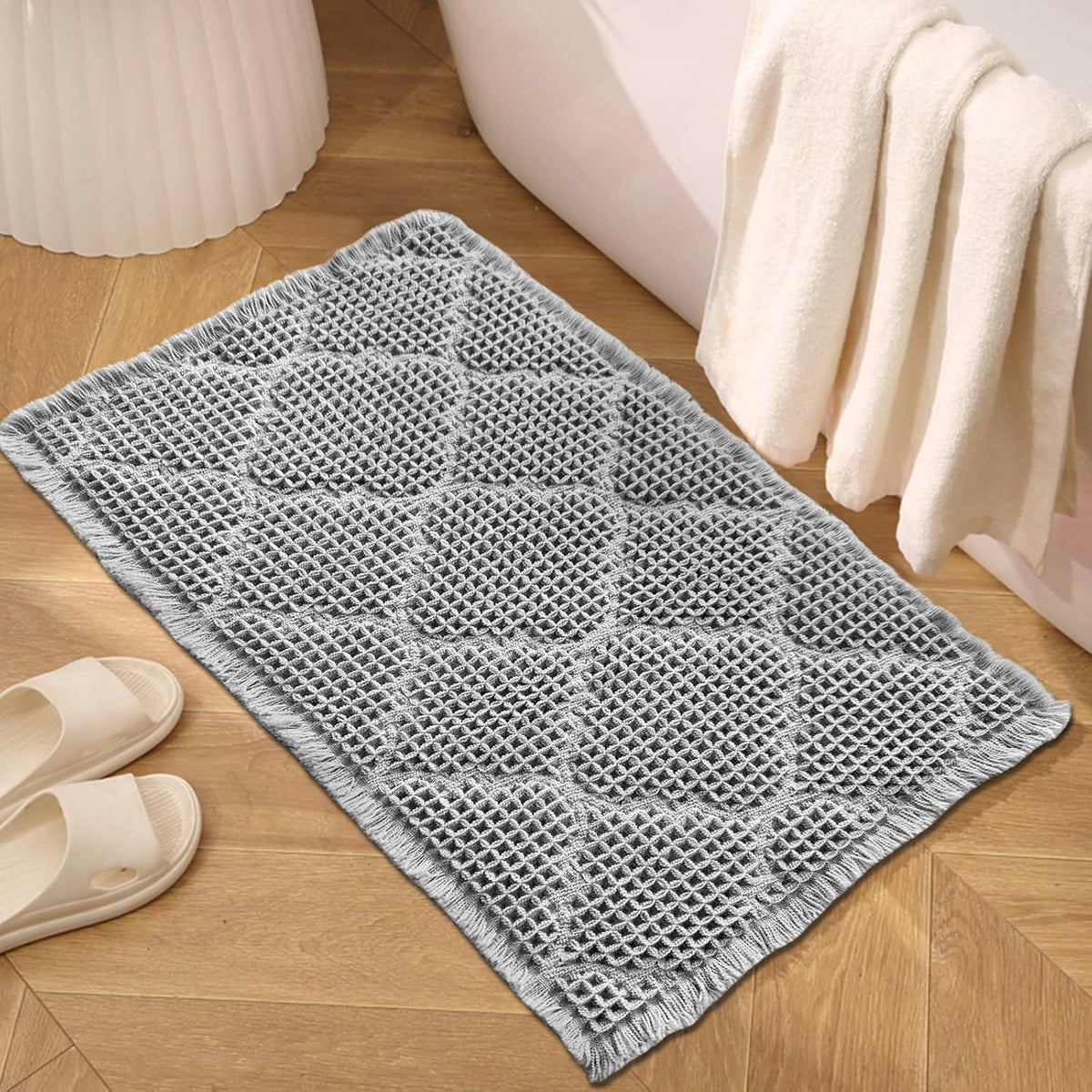Small waffle bath mat with fringe placed next to a bathtub and hanging towel on wood flooring