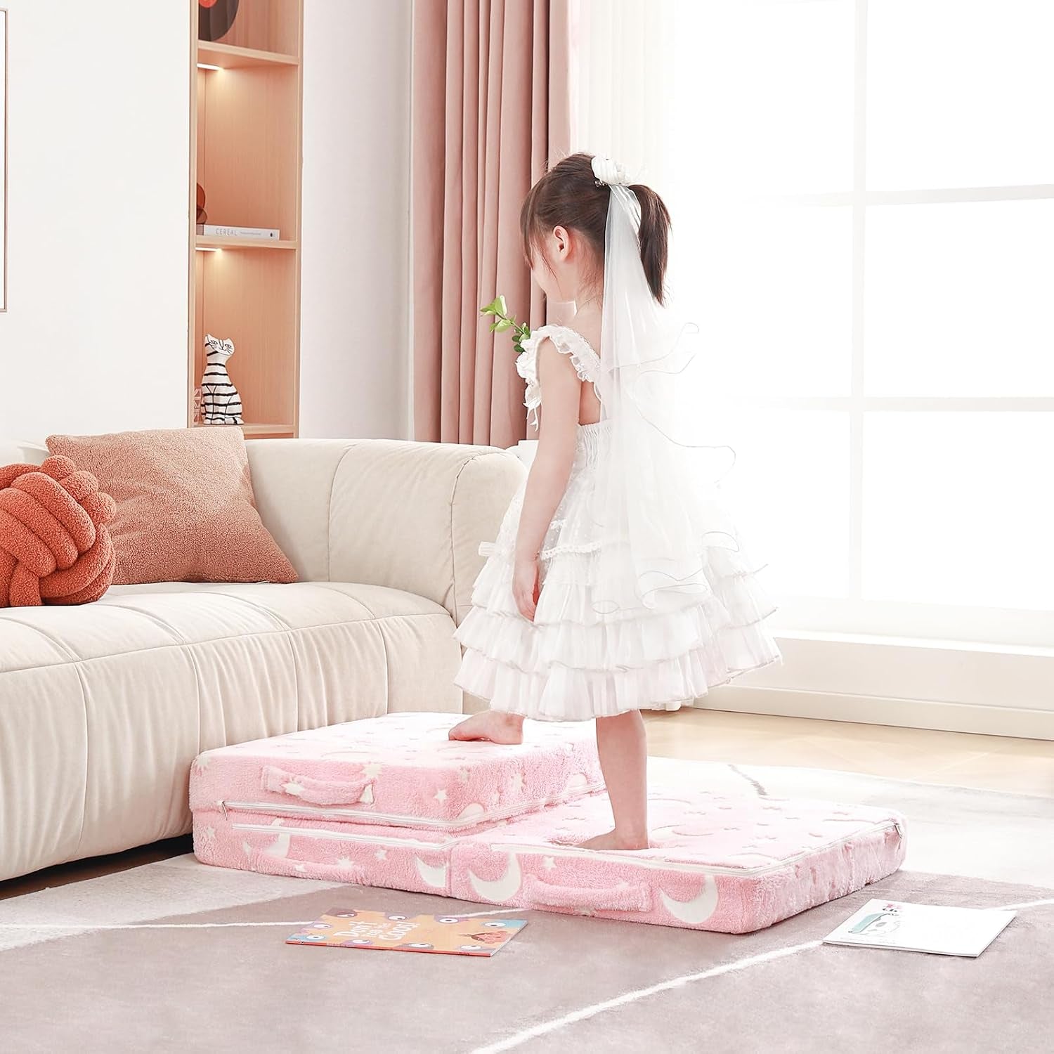 Child standing with one foot on the unfolded pink mattress laid on the floor in front of a sofa.