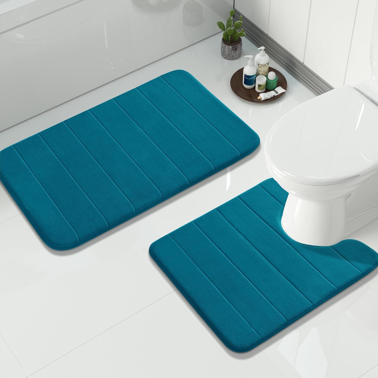 Two teal memory-foam bathroom mats on glossy white tile with toiletries and a toilet nearby