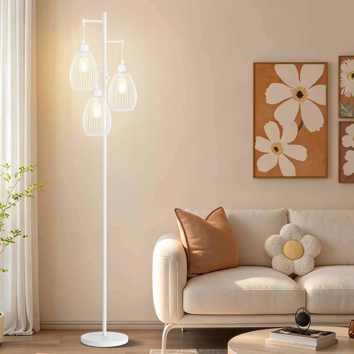 White three‑cage floor lamp glowing next to a cream sofa with floral wall art and a round coffee table.
