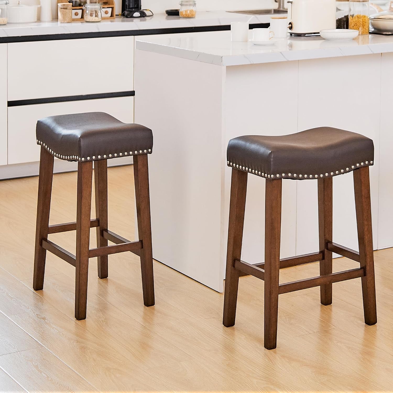 Two dark brown upholstered saddle stools with wooden legs beside a kitchen island on wood flooring.
