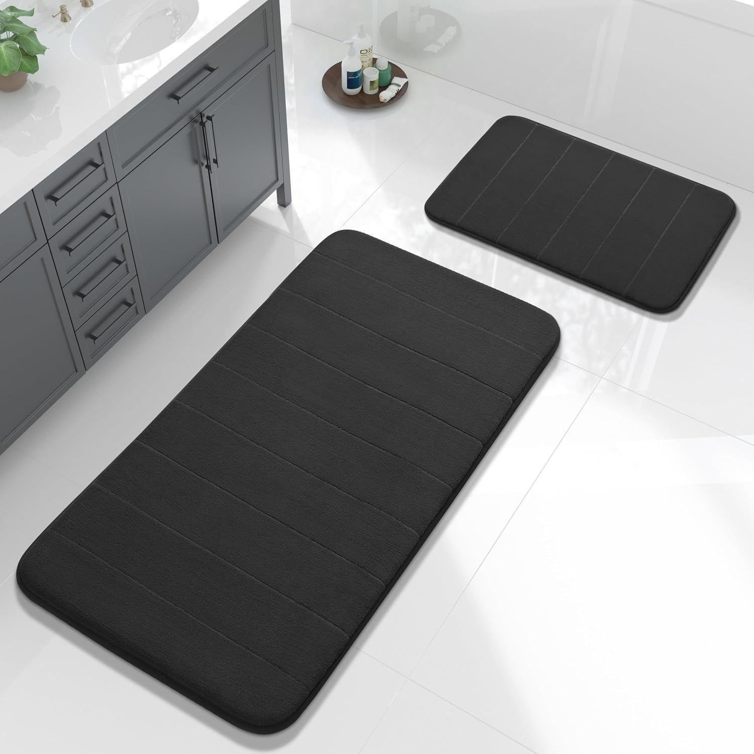 Black large rectangular mat and smaller toilet contour rug on white tile beside a gray vanity