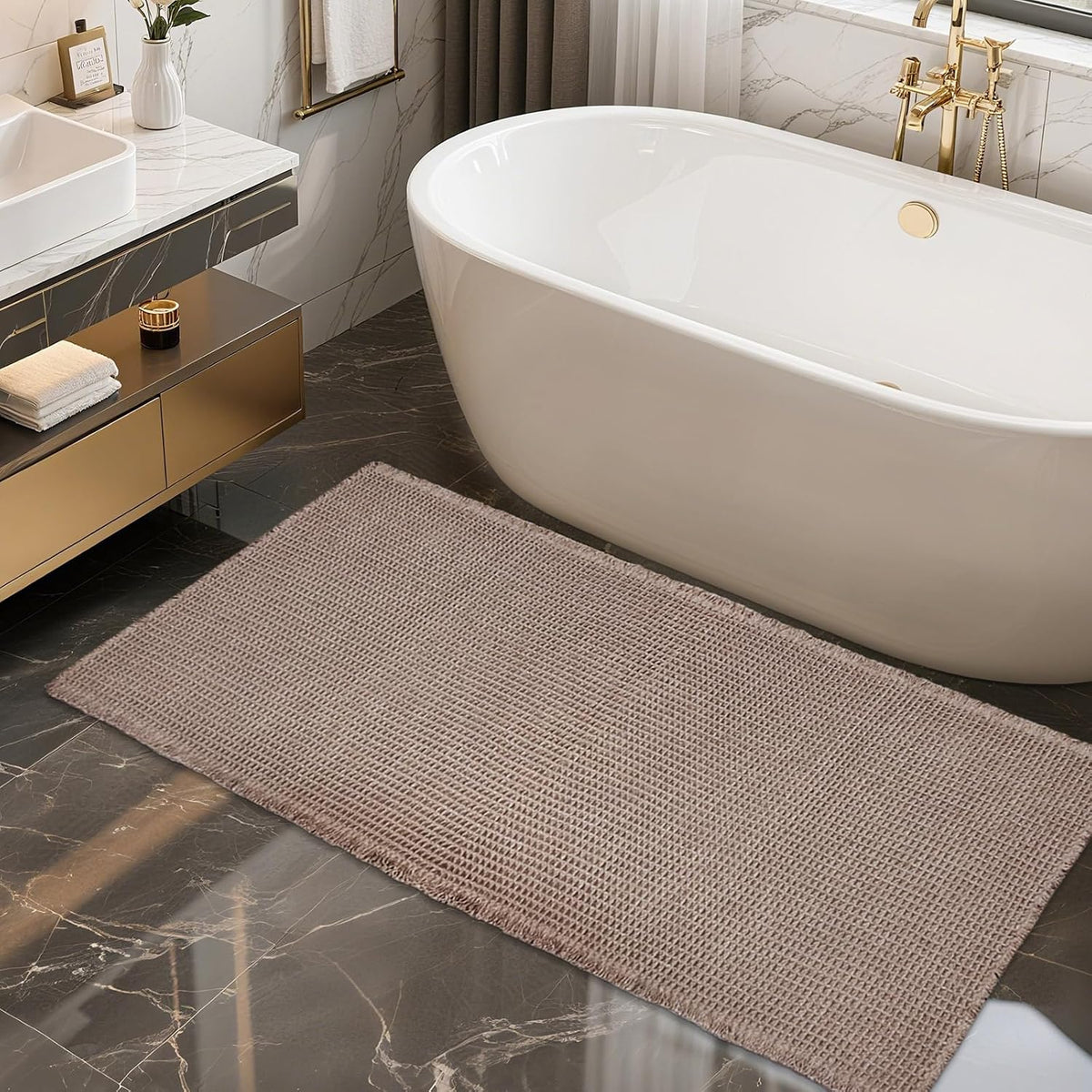 Low-angle side view of a taupe waffle bath mat beside a white freestanding tub and gold faucet.