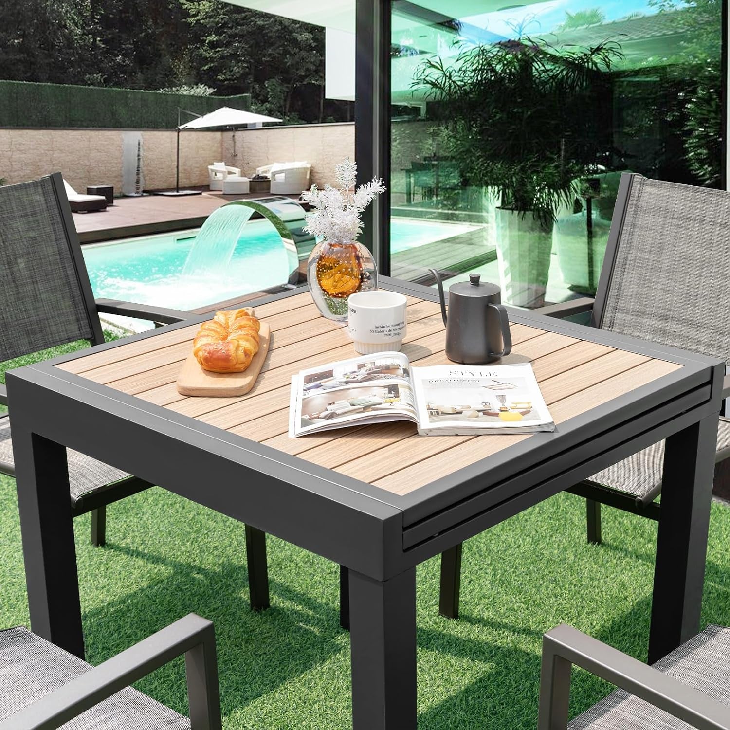 Aluminum patio table in dark gray with wood-like slatted square top shown on lawn near a pool with two outdoor chairs
