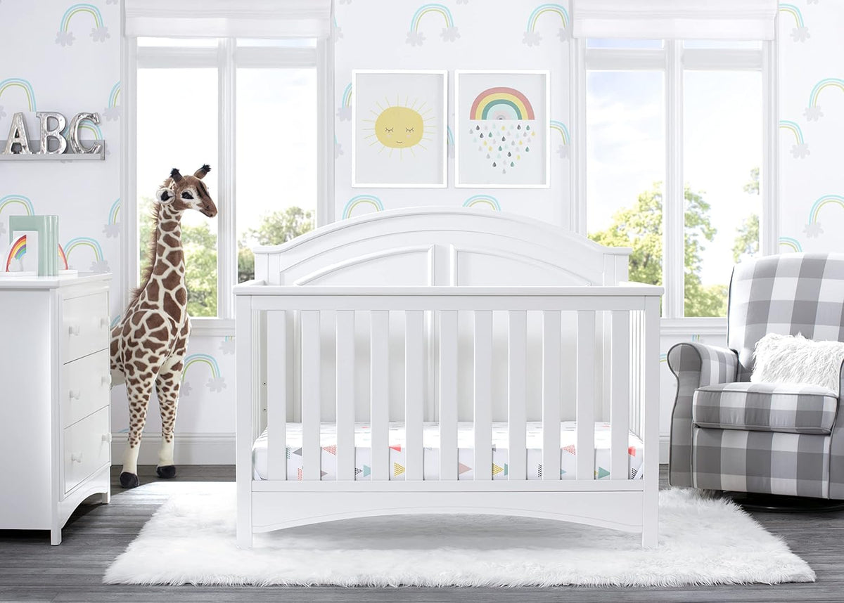 Front-facing crib staged in a bright nursery with rug, dresser, stuffed giraffe, and armchair.