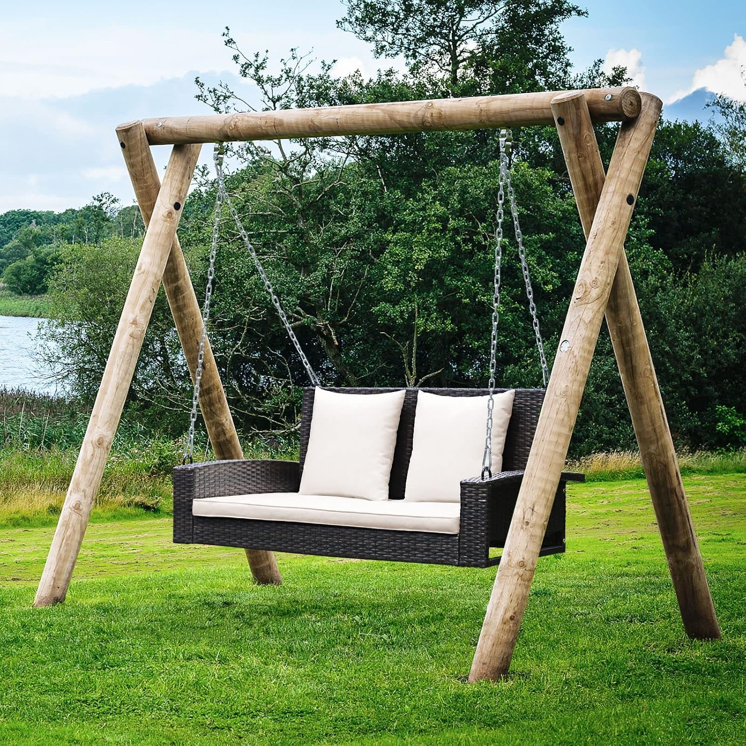 Dark brown rattan hanging porch swing with cream cushions suspended from a wooden A-frame in a grassy lakeside setting