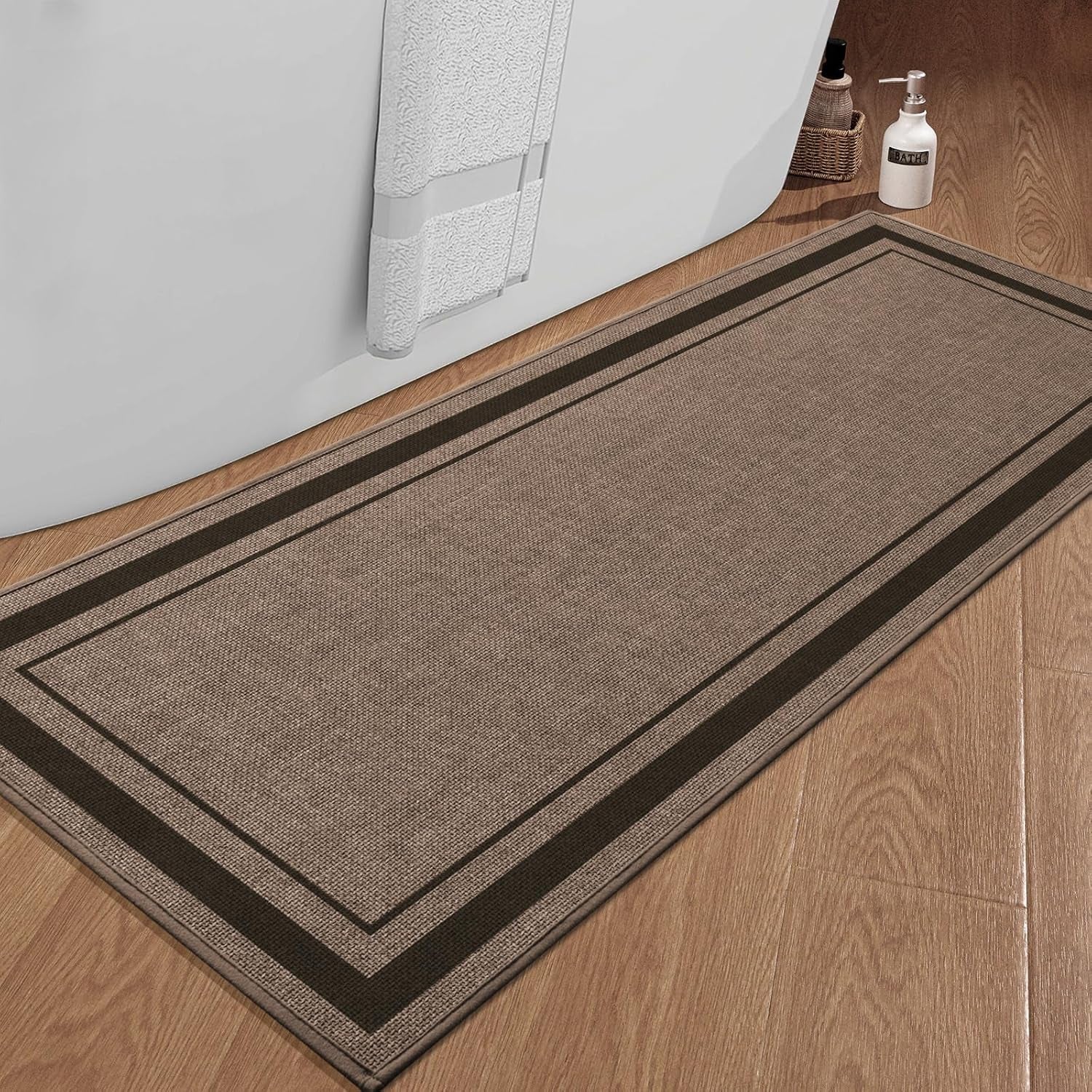 Brown rectangular bath rug with dark twin borders placed beside a white tub on wood flooring