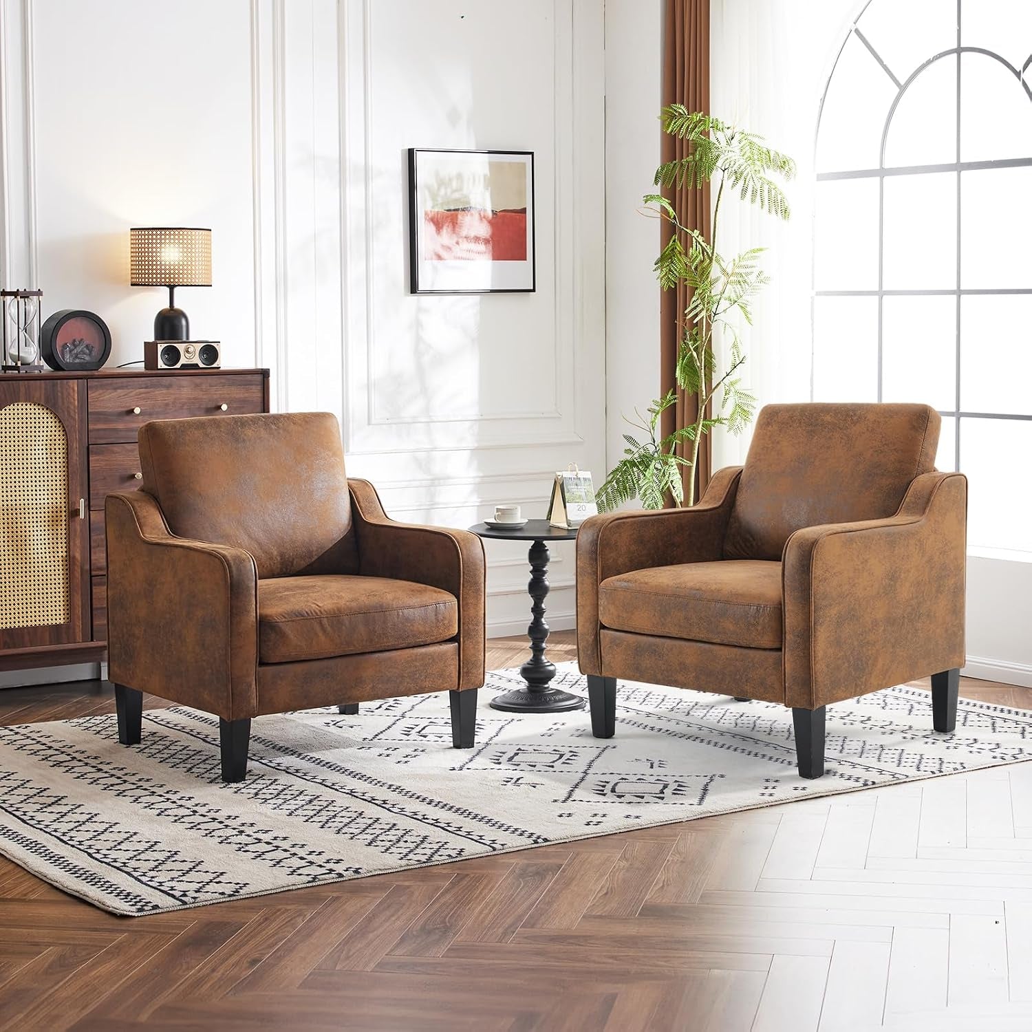Pair of brown mid-century upholstered accent chairs with black legs flanking a small round black side table in a sunlit