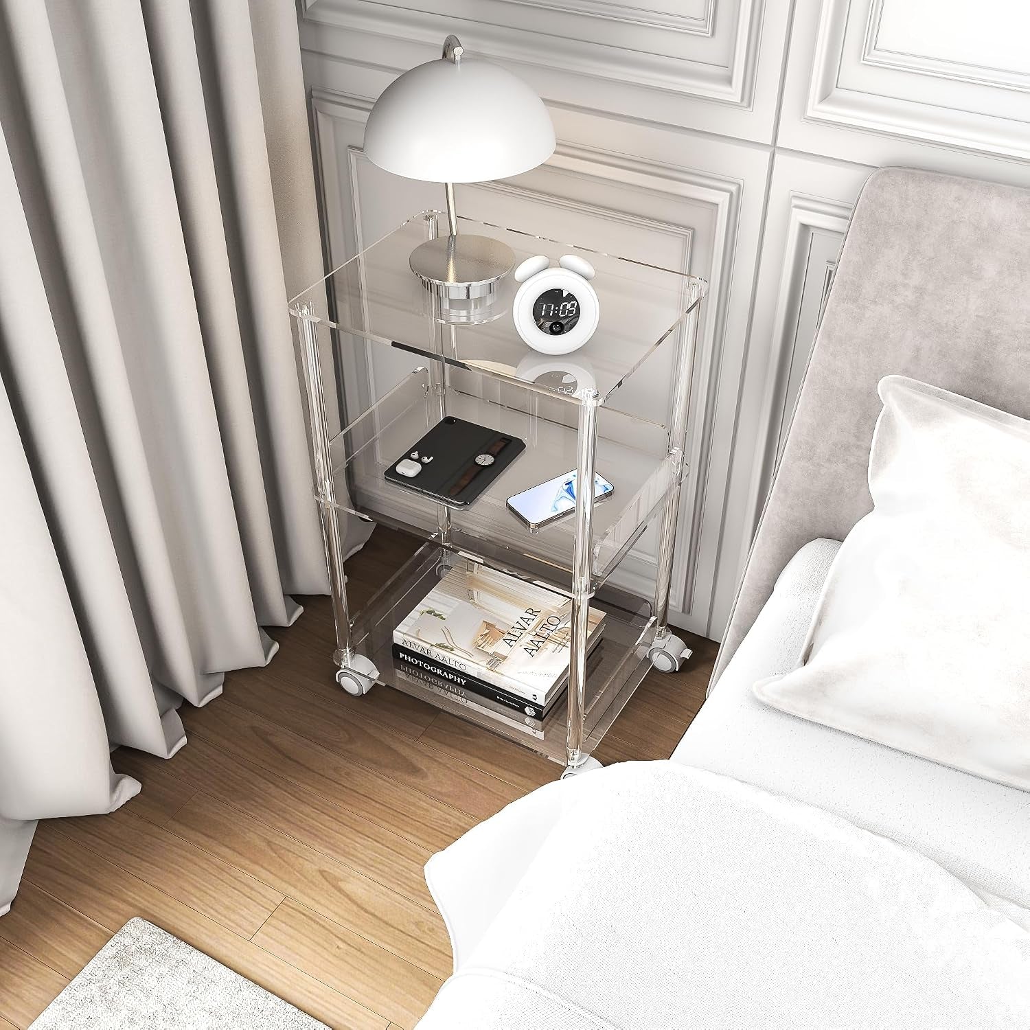 Clear 3-tier acrylic stand used as a bedside table with lamp, alarm clock, phone, and books.