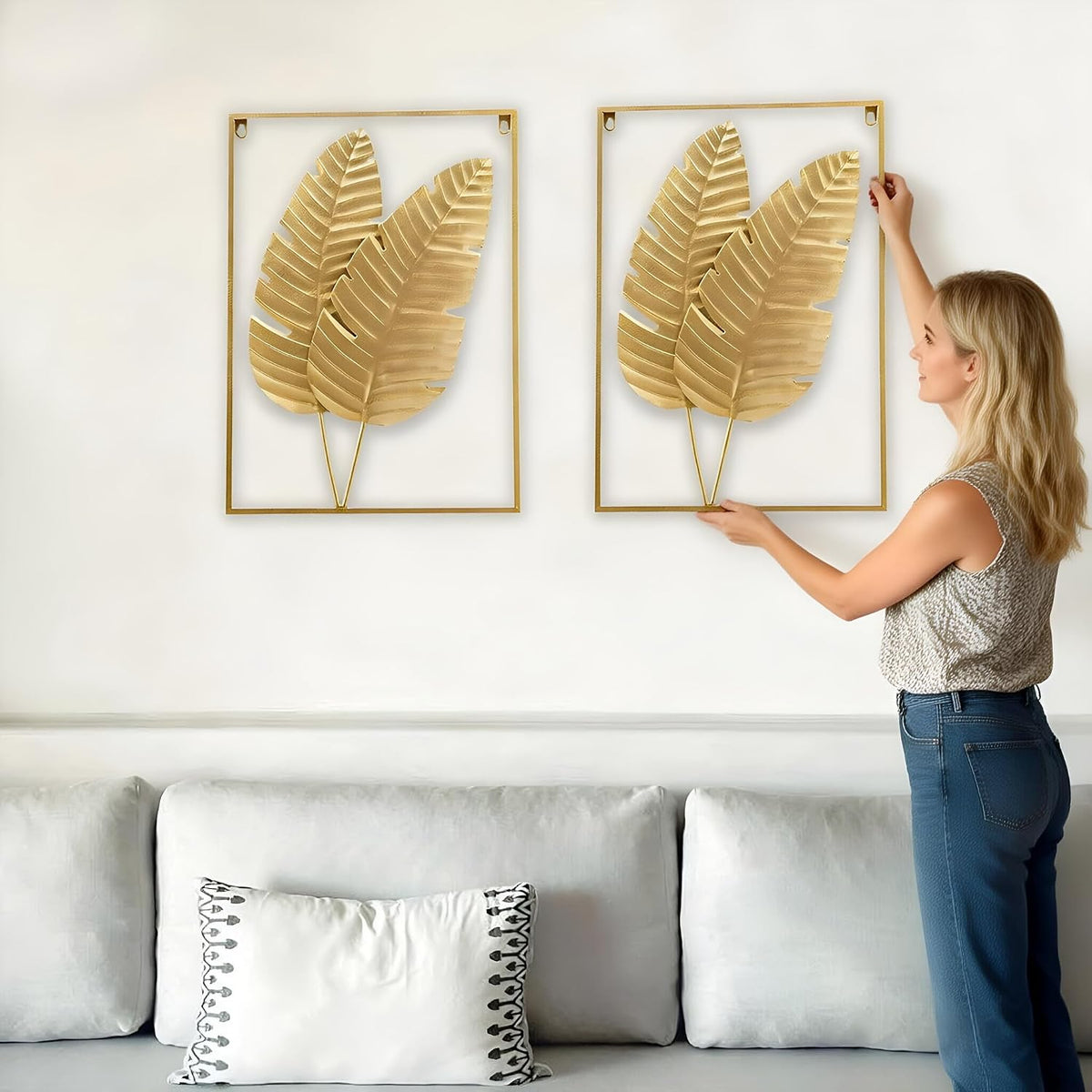 Woman holding one gold metal leaf frame while placing the pair above a light grey sofa