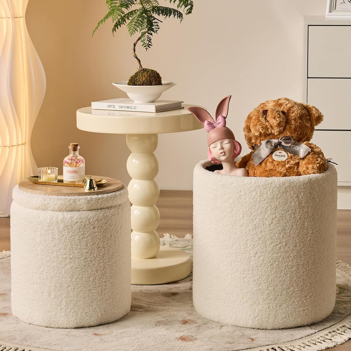 Cream textured ottoman pair, one open showing stuffed bear inside beside a glossy cream side table with bonsai.