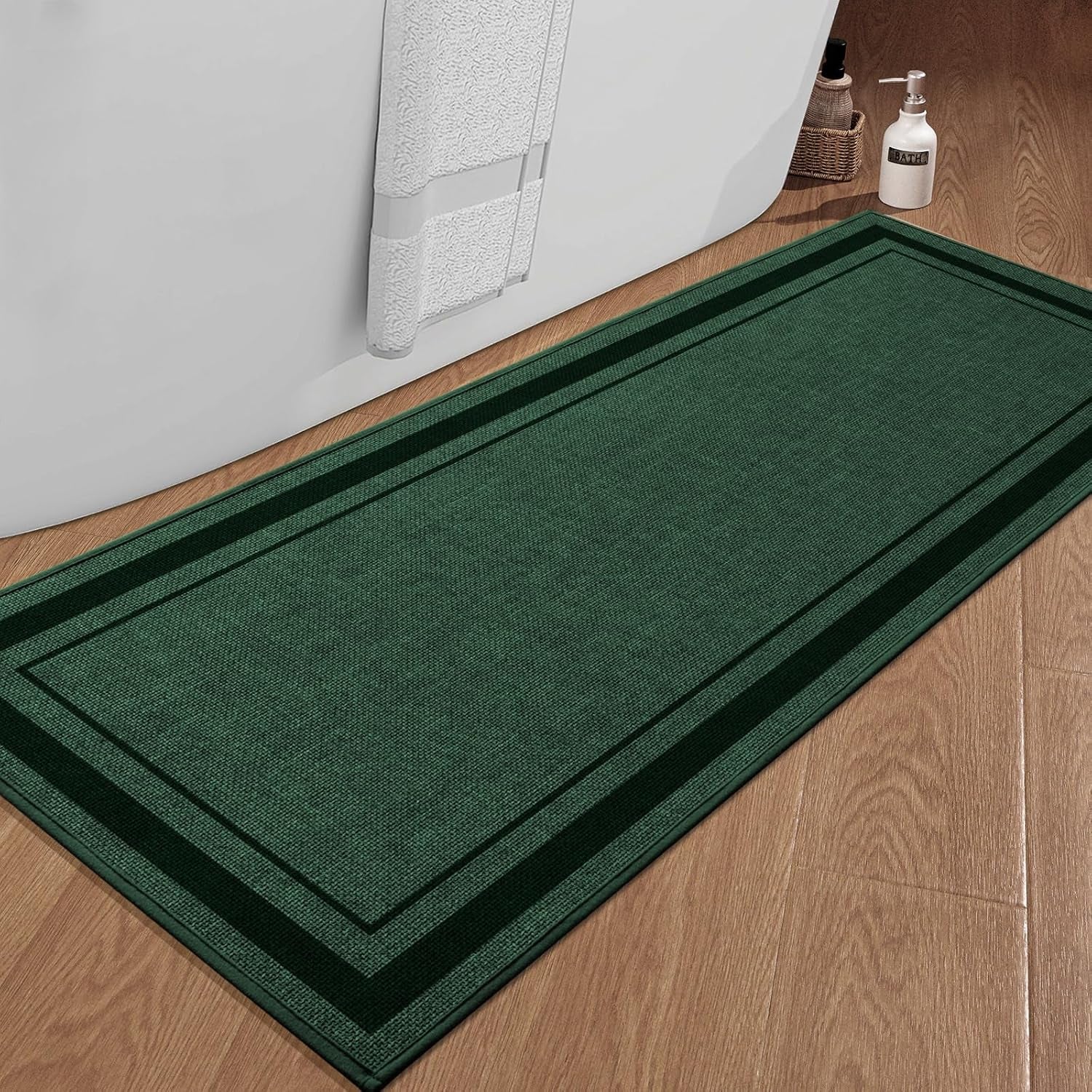 Dark green rectangular bath rug on wood floor beside a freestanding tub with towel and soap bottle