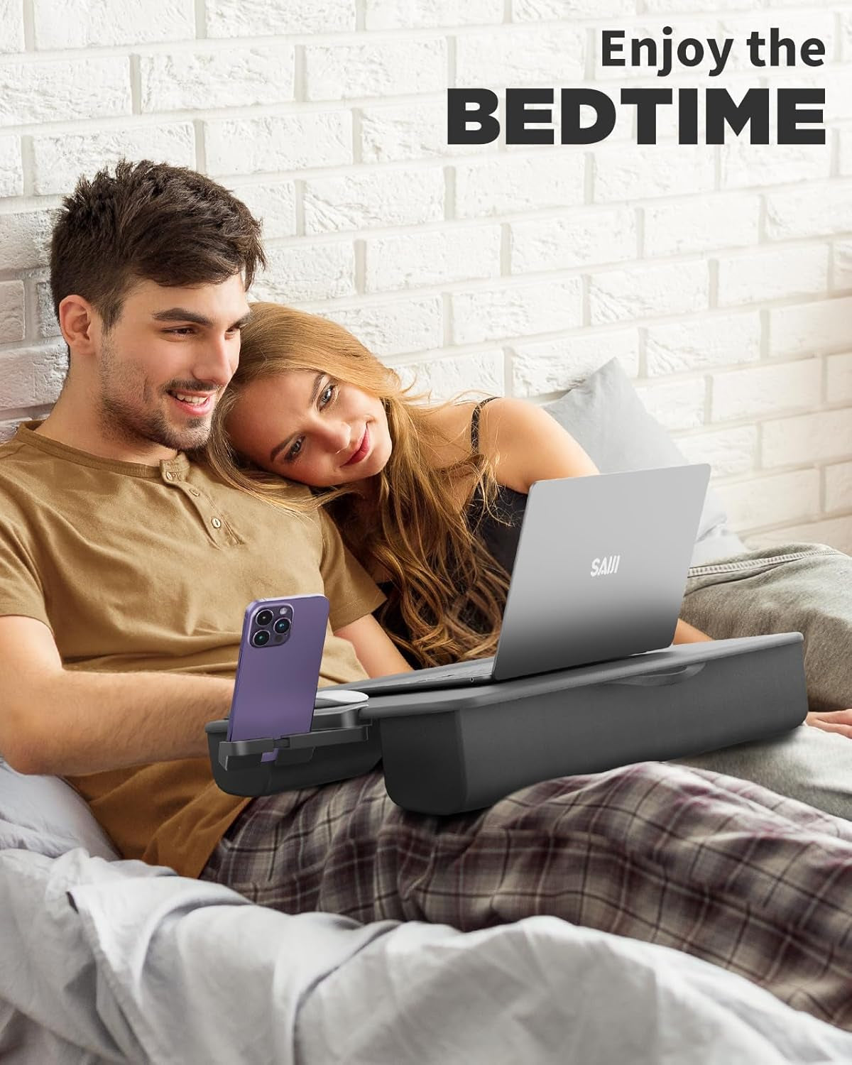 A couple reclining in bed sharing a lap desk that holds a laptop and a phone in its built-in holder.
