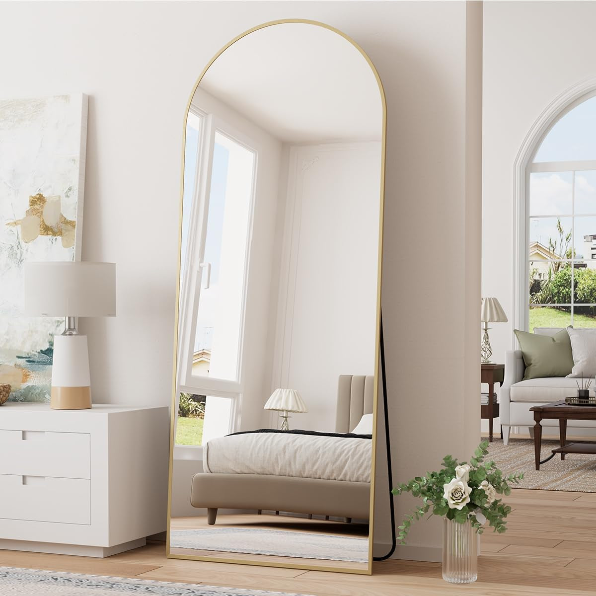 Gold‑framed arched floor mirror leaning against a wall reflecting a bed and window in a bright bedroom.