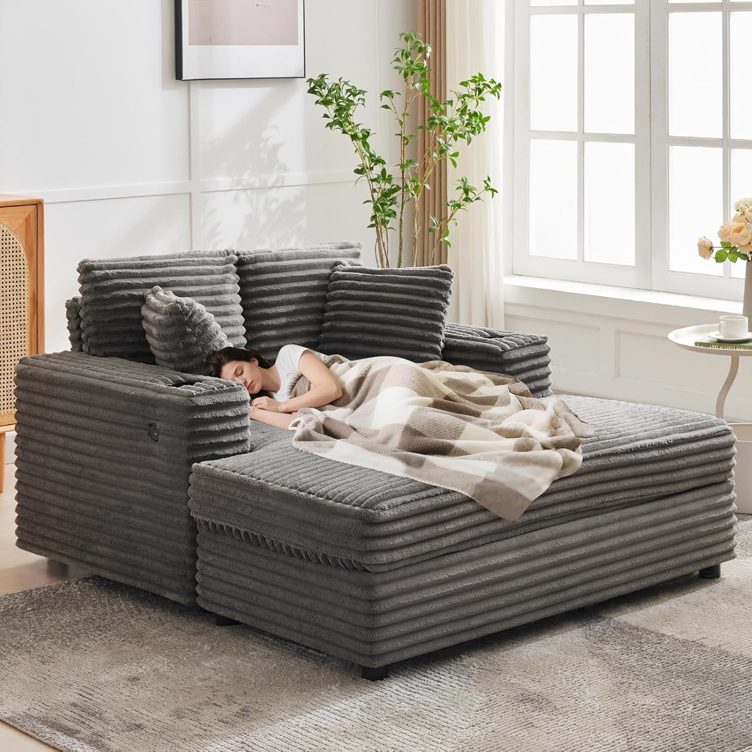 Angled side view of the gray corduroy chaise in a bright room with a woman sleeping under a checked blanket.