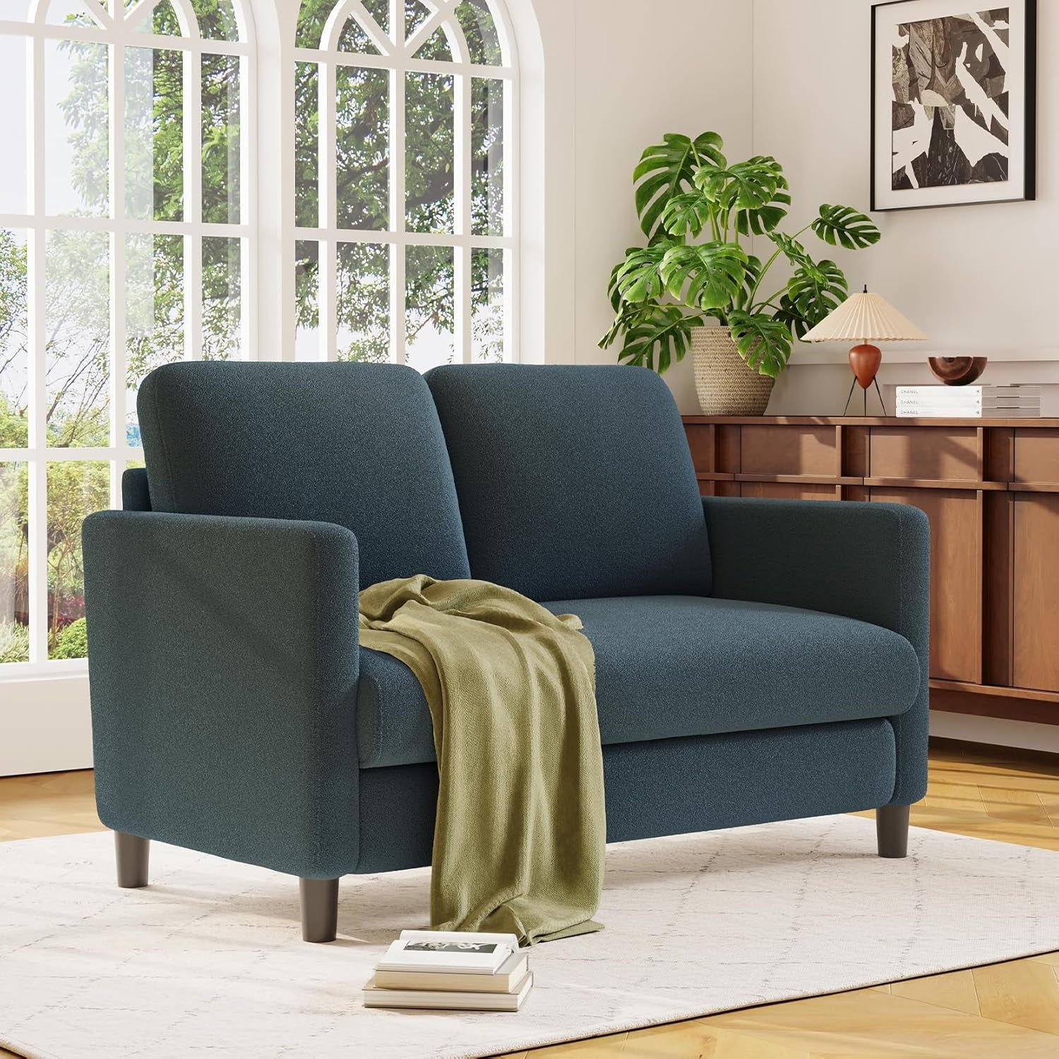 Three-quarter view of a blue-gray loveseat with a green throw draped over the seat, books on the floor and a wooden console