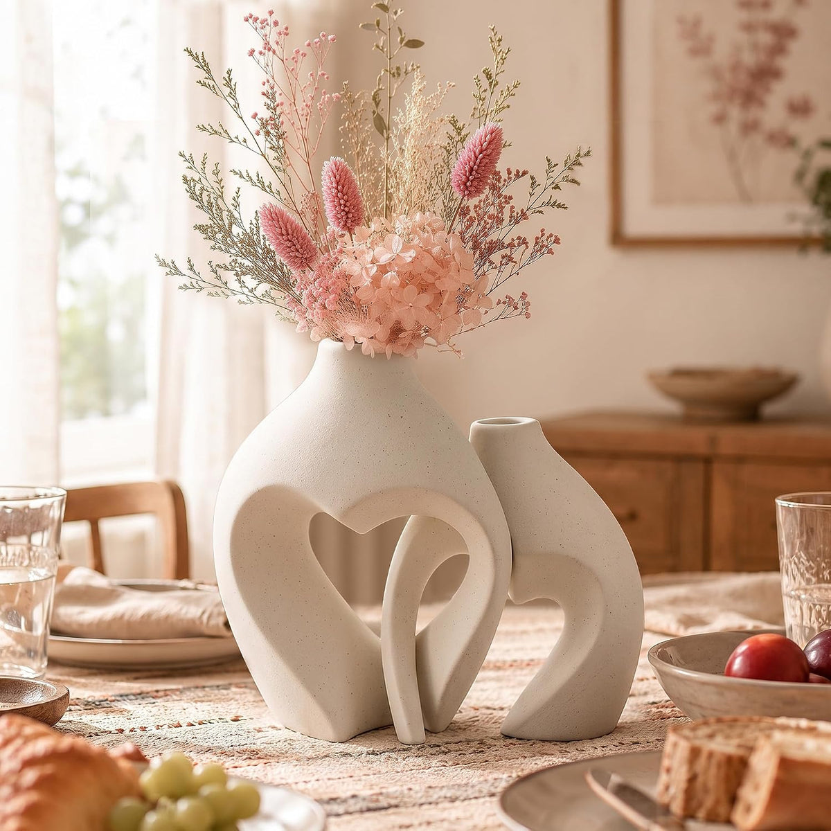 Two sand-colored heart vases on a rustic breakfast table surrounded by bread, grapes, and dishes