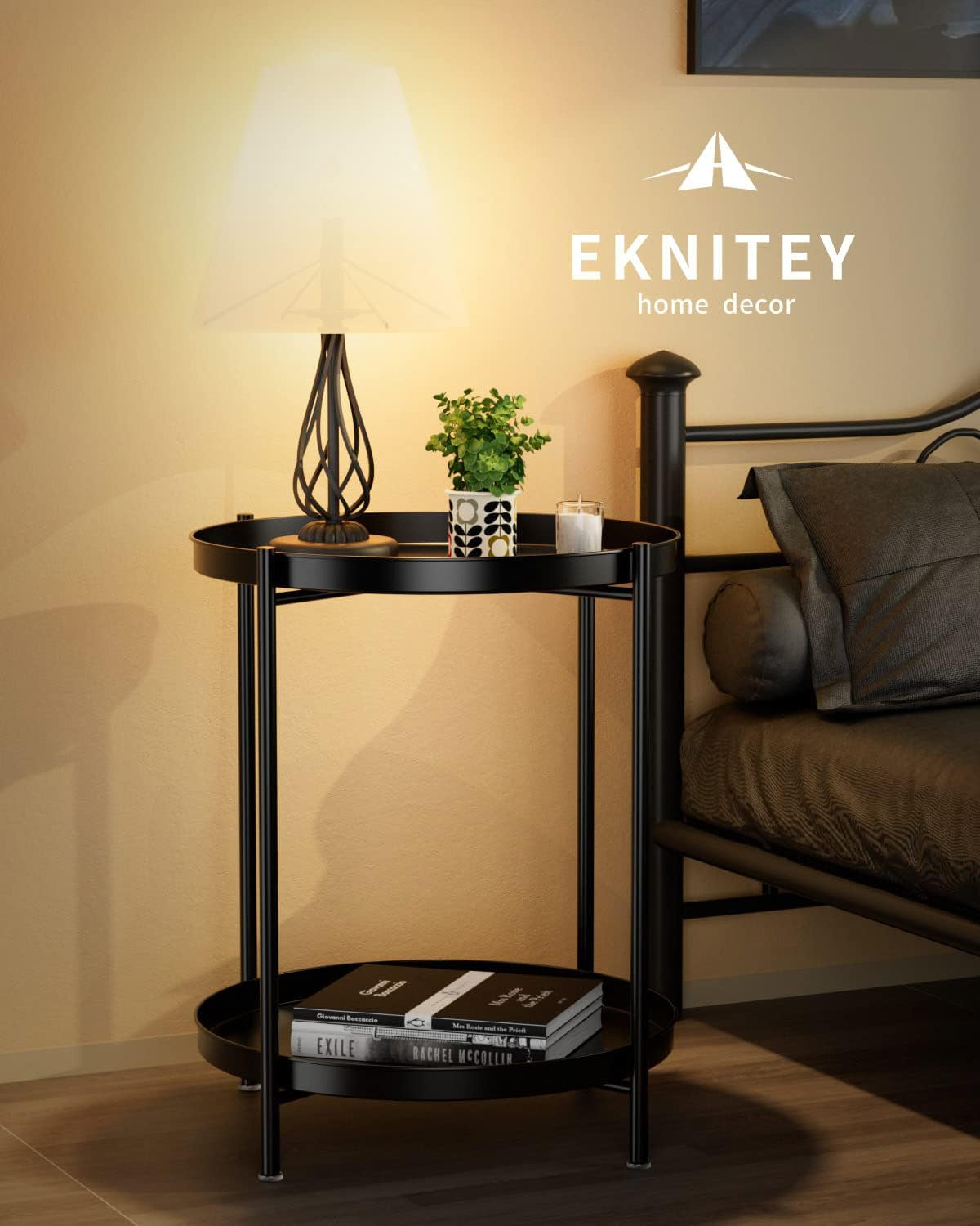 Black round metal end table next to a bed with lamp, plant and books, brand logo visible on wall.