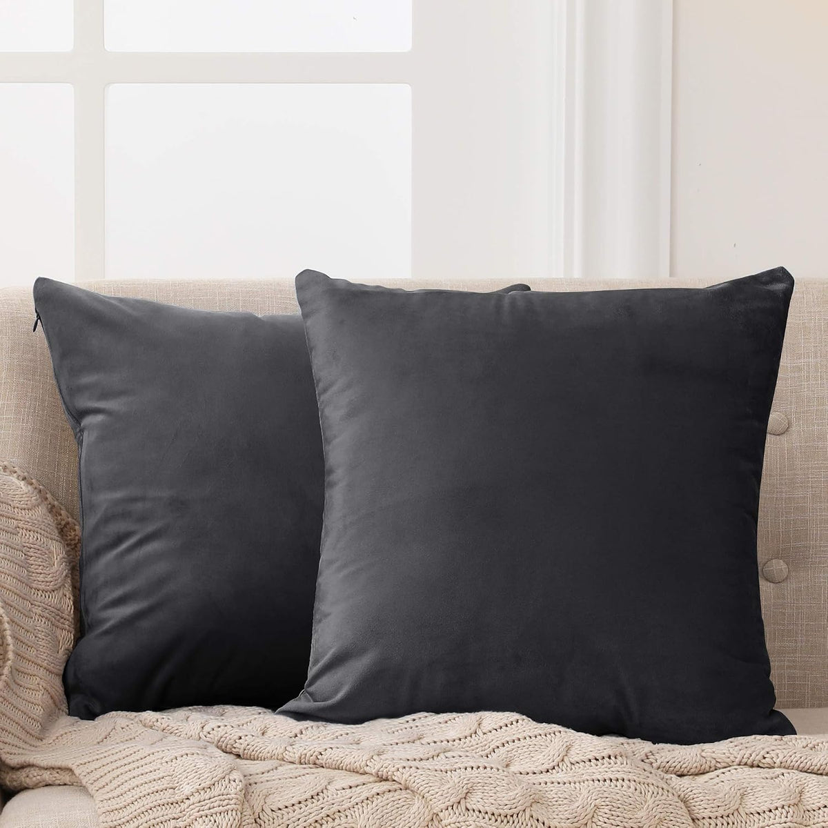 Close-up of two charcoal dark-gray square pillow covers on a beige couch with a knitted throw.