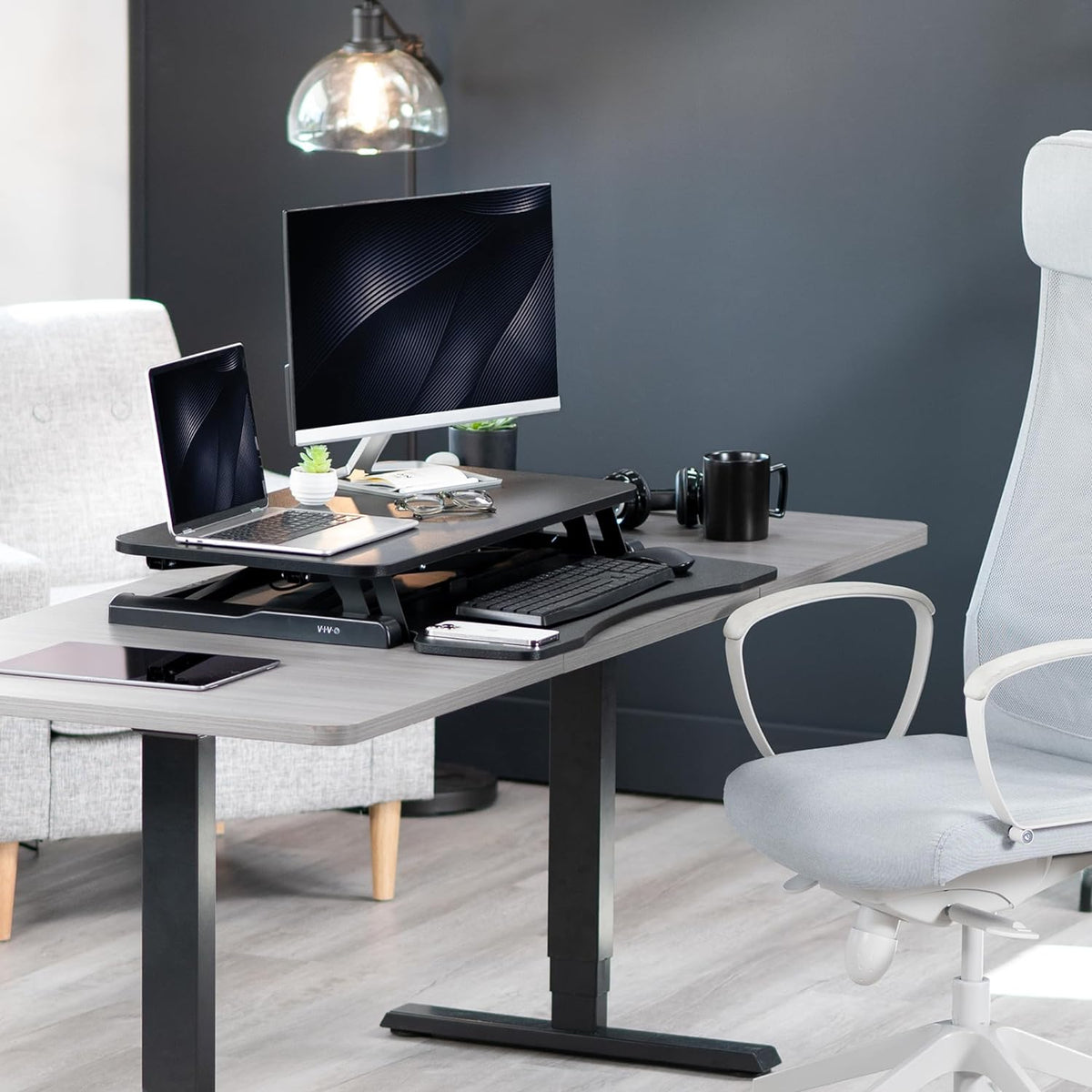 Black standing desk converter on a grey desk with monitor, laptop, mug and a light grey office chair in a home office
