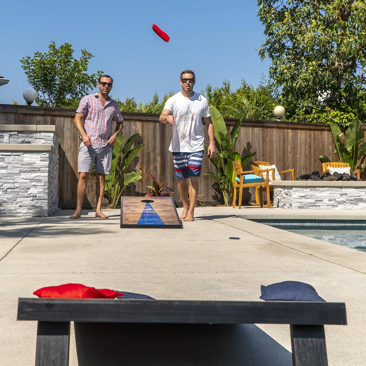 Poolside action shot showing a red bag in midair and two men throwing toward a barnwood-style board.