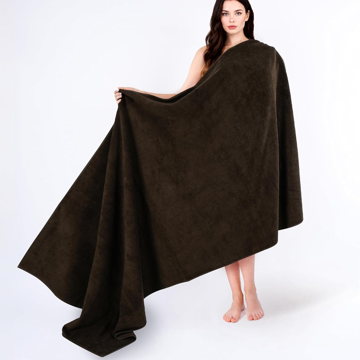 Woman standing barefoot holding and wrapped in a very large dark brown bath towel against white background.