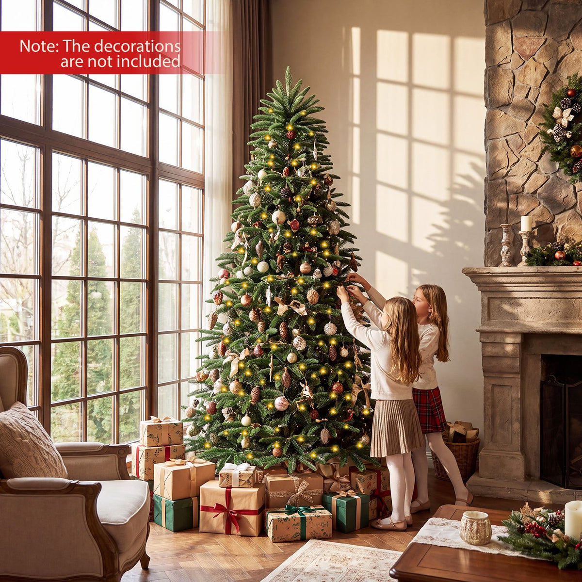 Two girls decorate a glowing Christmas tree surrounded by presents near a living room fireplace.