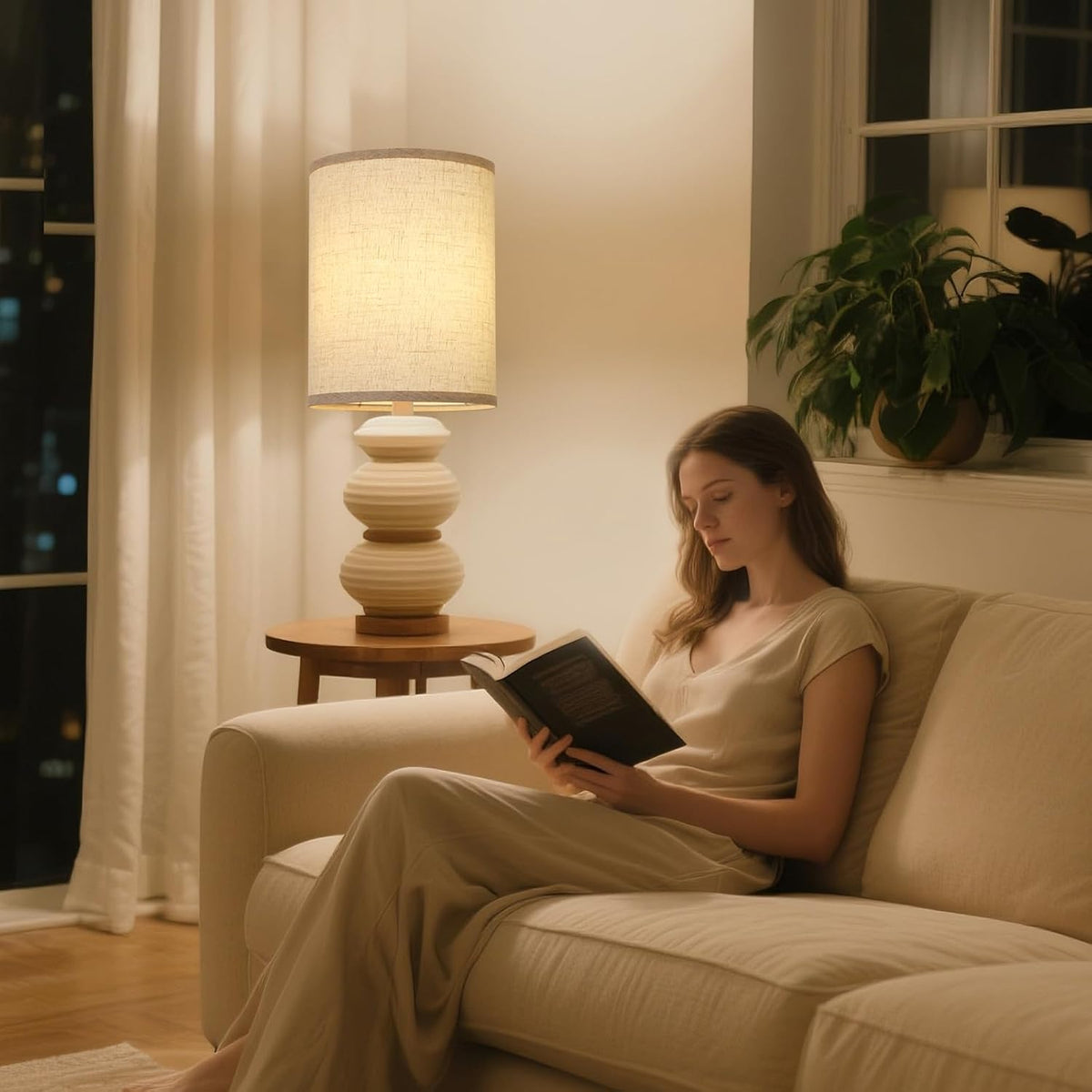 Lit beige drum lampshade on table lamp casting warm, diffused light over a woman reading on sofa
