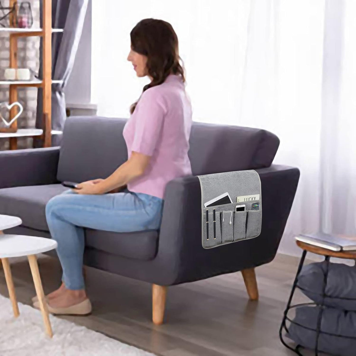 Grey armrest organizer draped over a dark sofa arm in a living room beside a seated woman.