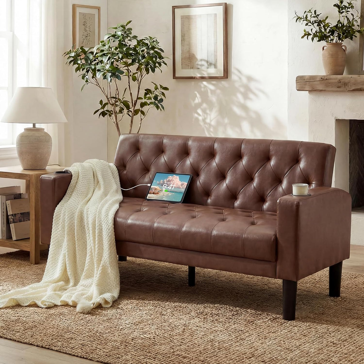 Warm brown tufted loveseat shown from the side with a charging tablet on the seat and a chunky cream throw.