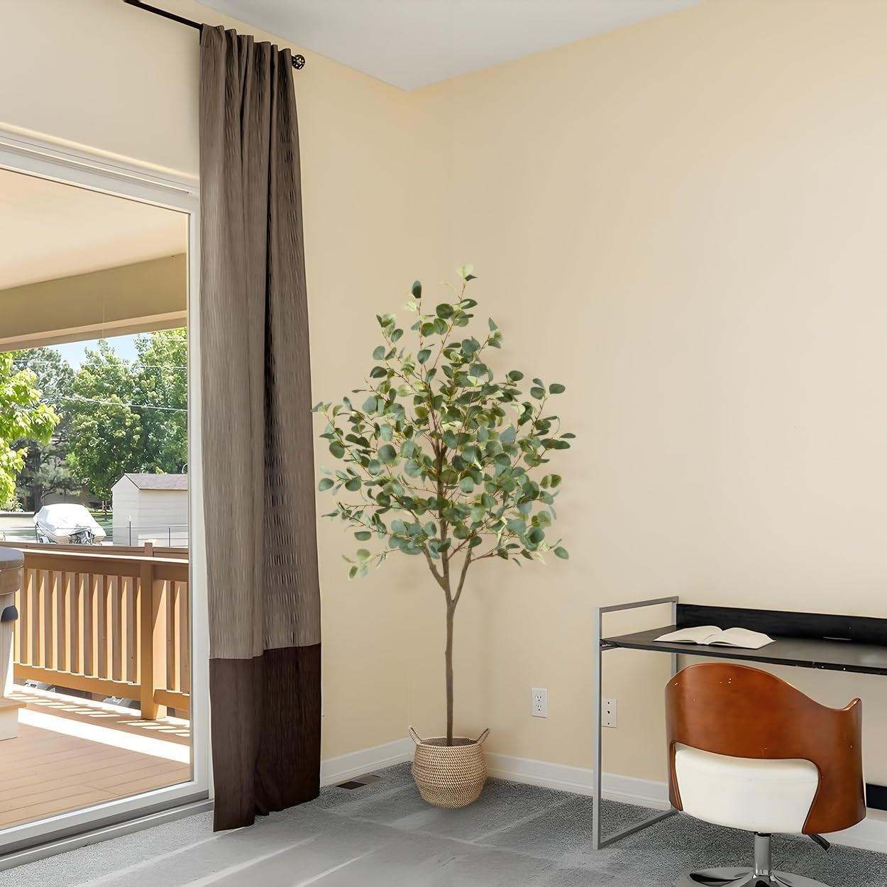 Corner of a bright room with a tall artificial eucalyptus tree in a woven basket beside a desk and sliding door.