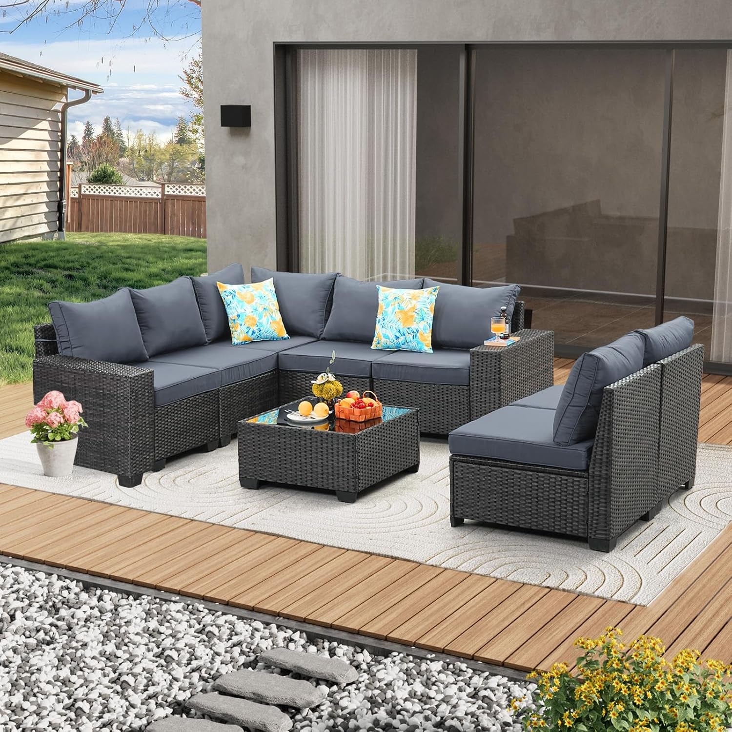 Outdoor corner rattan sectional with dark gray cushions, glass-top coffee table and floral throw pillows on a deck.