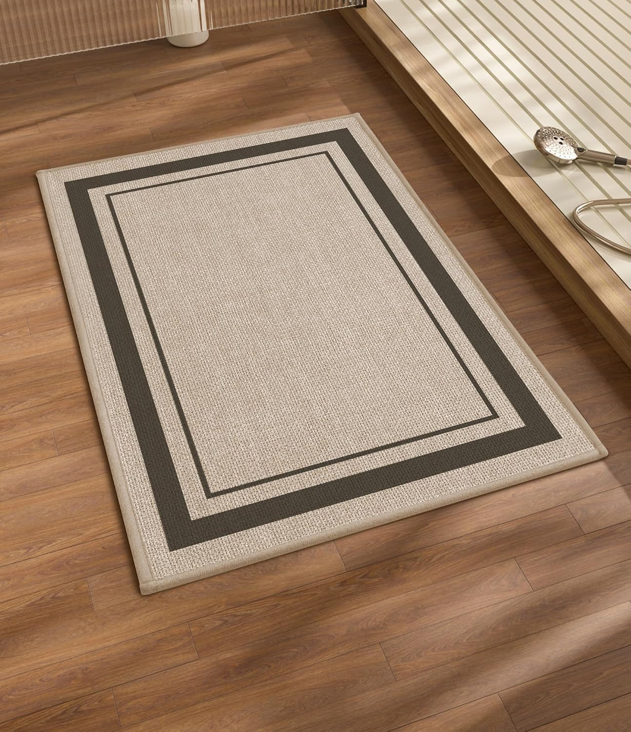 Rectangular beige bath mat with double dark border laid on wood floor beside a raised tub and handheld showerhead.