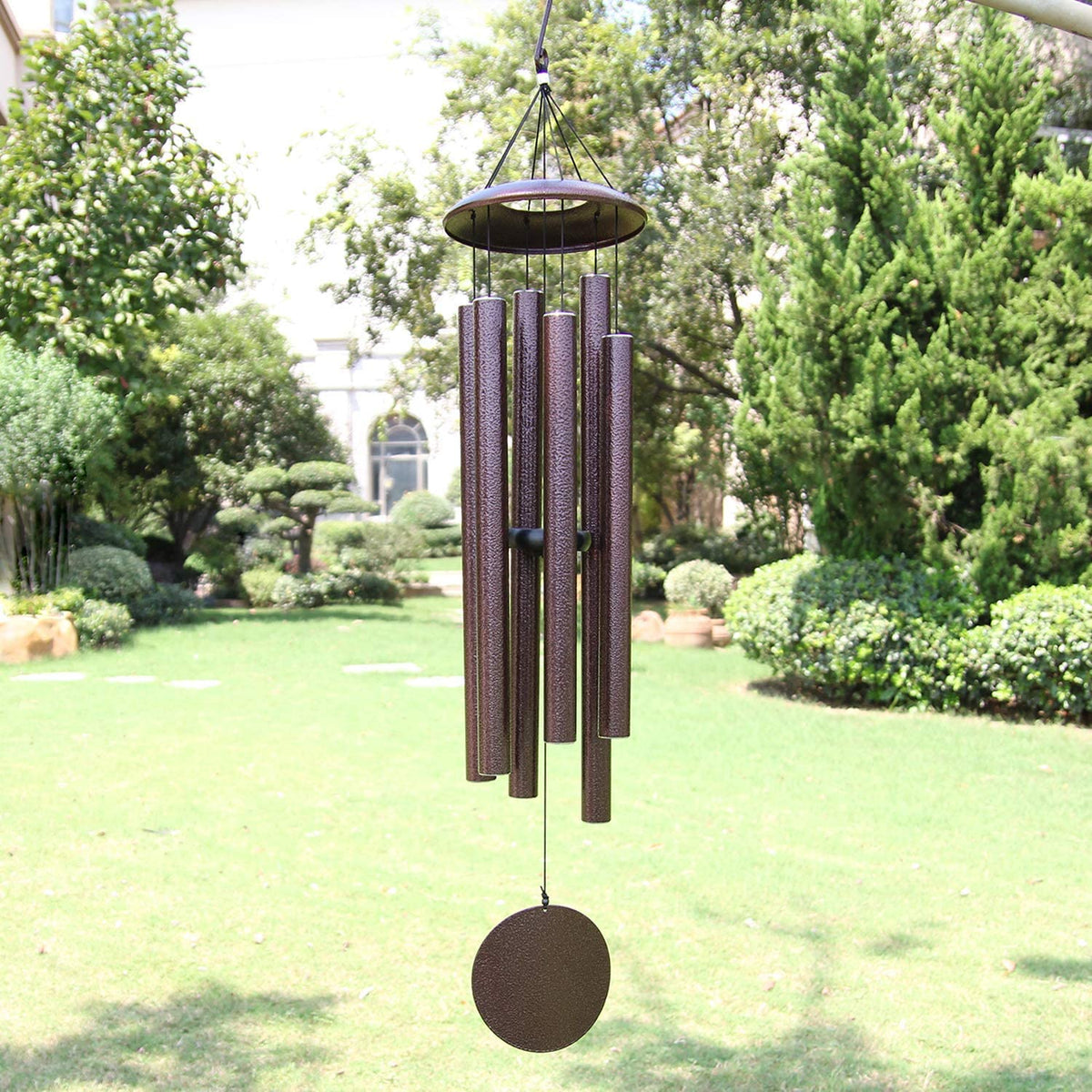 Outdoor scene showing a hanging brown wind chime with six metal tubes and round sail.