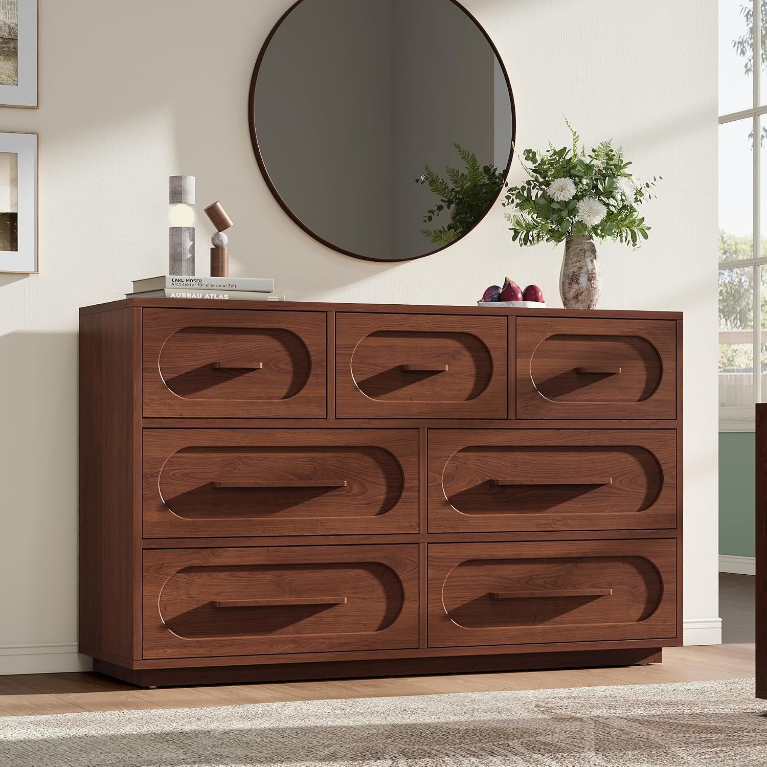 Wide dark-brown multi-drawer dresser under a round wall mirror with vase and books on top