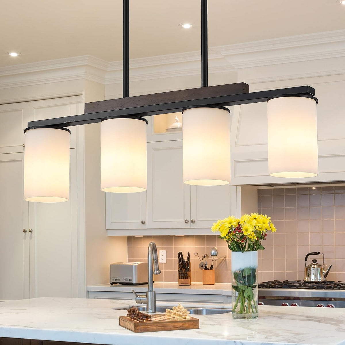 Kitchen island view with four lit white cylindrical shades on a black linear chandelier above sink, vase of yellow flowers