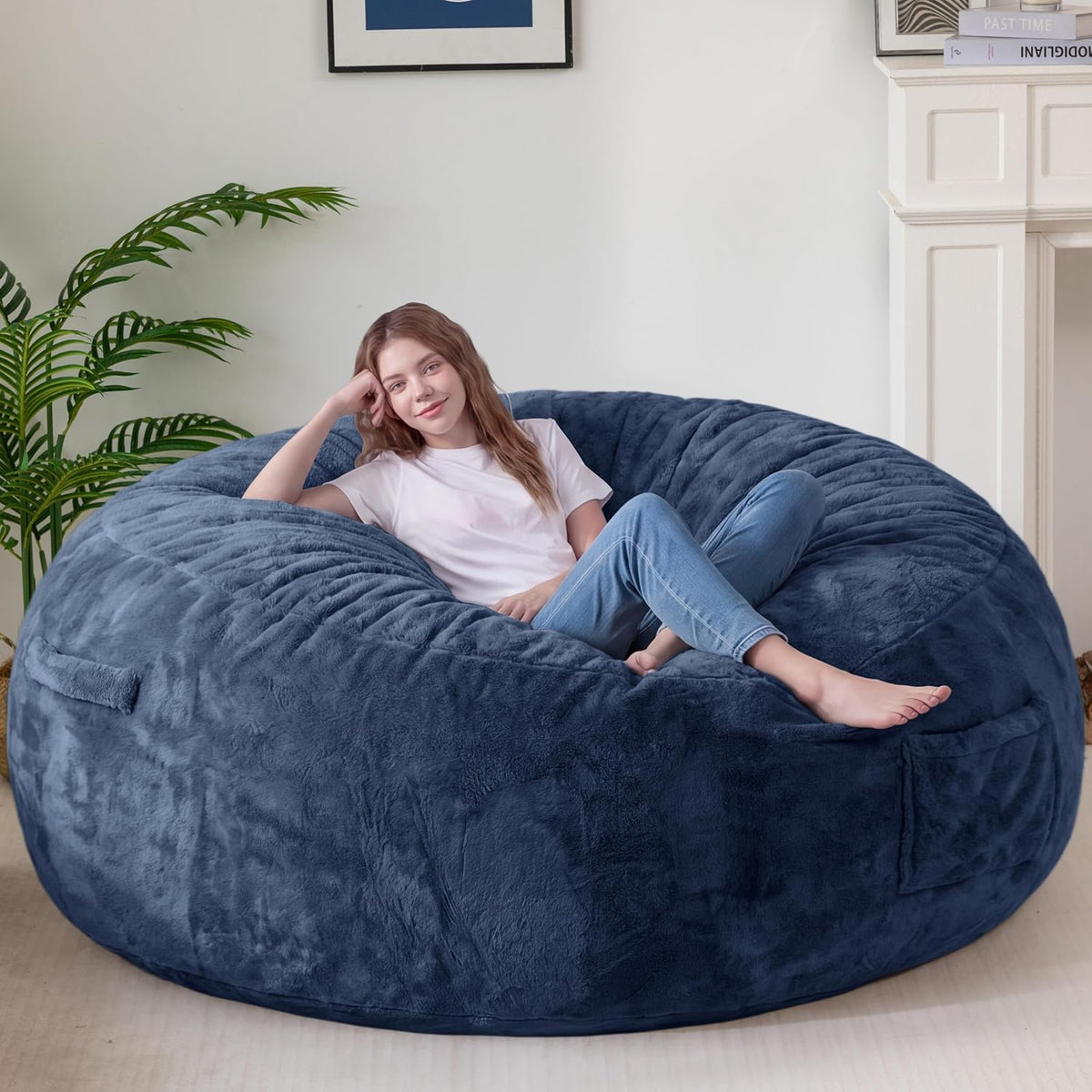 Angled lifestyle shot of a navy faux‑fur bean bag with a woman reclining, plant and mantel behind.