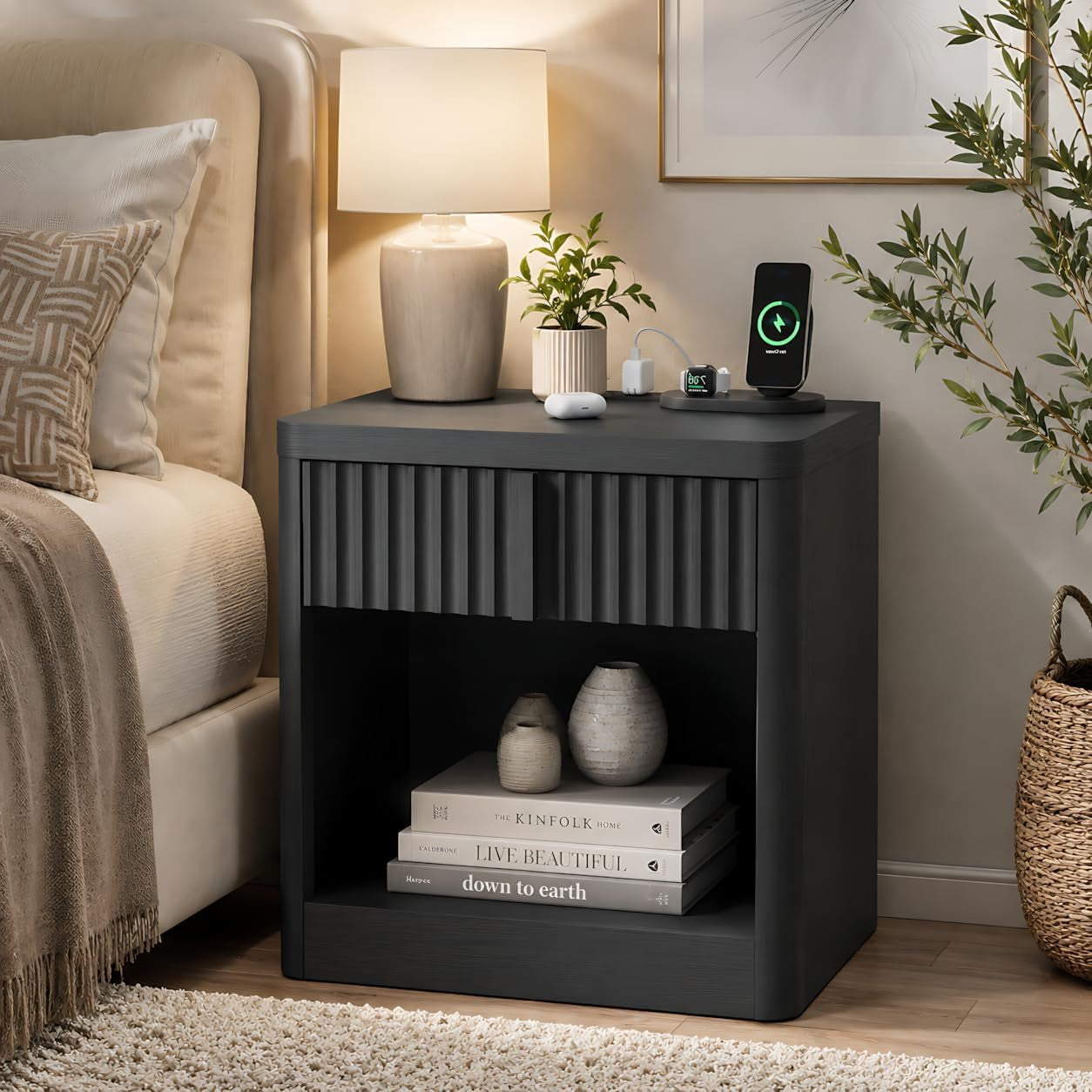 Black/dark fluted nightstand beside a bed, topped with a lamp and charging devices, open shelf holding books and ceramics.