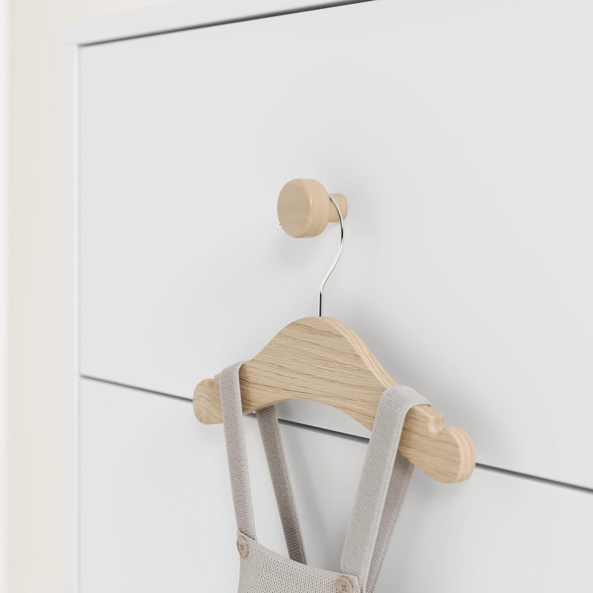 Close-up of a light wood round drawer knob on a white drawer with a small wooden hanger holding baby overalls.
