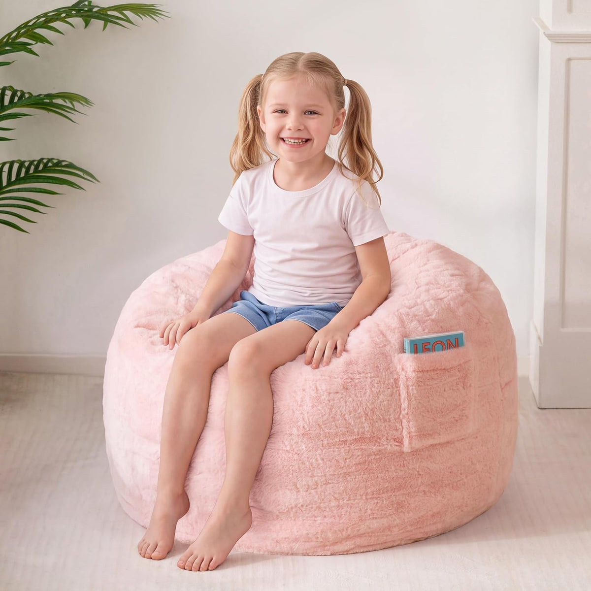Front view of a smiling young girl seated on a pale‑pink bean bag with a book visible in the side pocket.