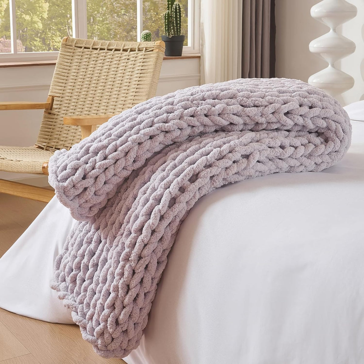 Light lavender chunky knit throw rolled and folded on the edge of a white bed beside a woven chair.