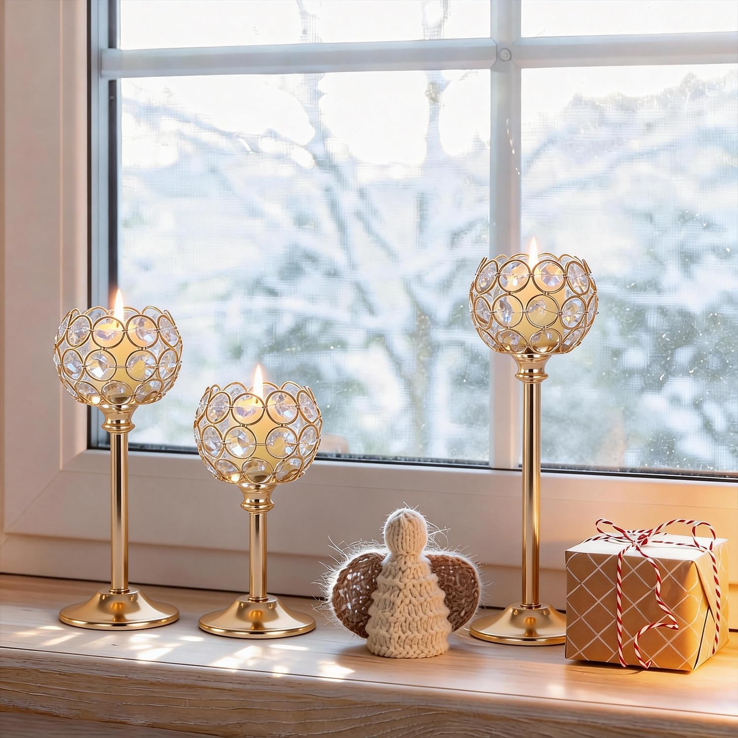 Three gold crystal holders lit on a windowsill with a snowy yard visible outside and a small wrapped gift nearby.