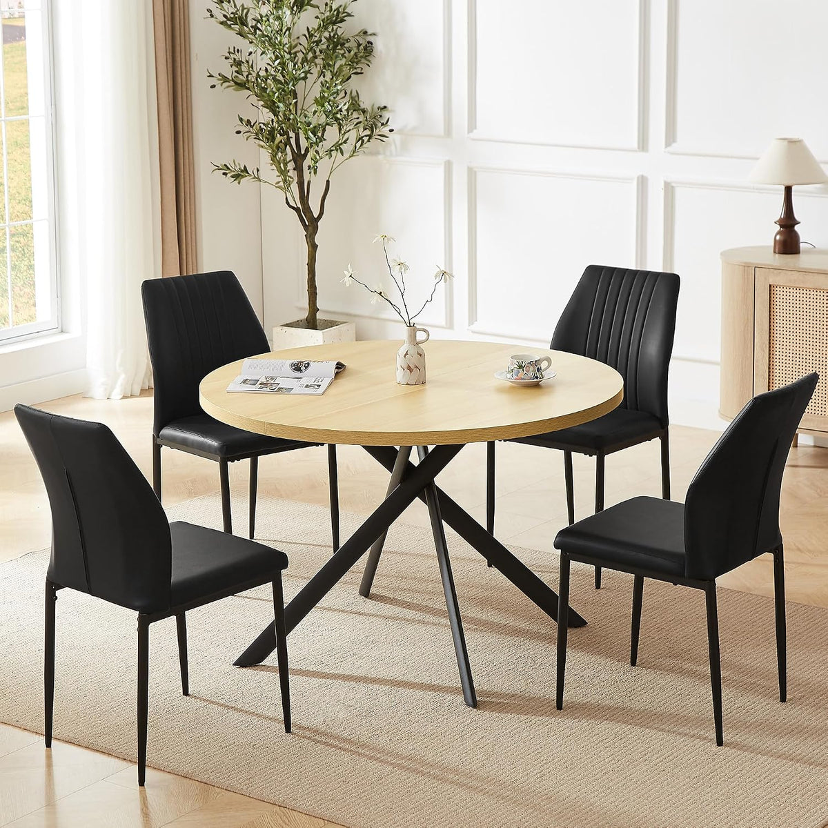 Round light-wood table with black U-shaped chairs in a bright dining room scene.