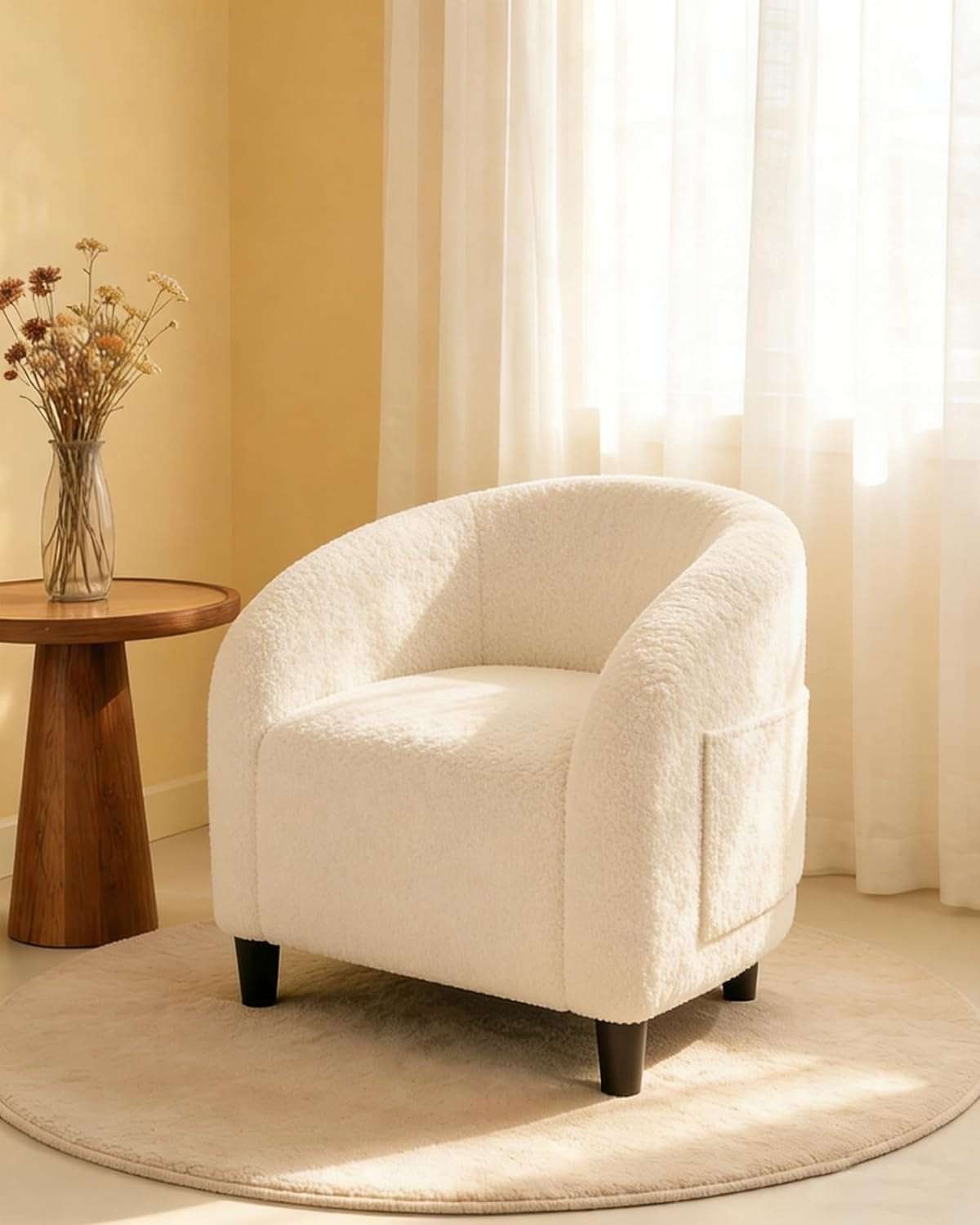 Single cream boucle barrel chair on a round beige rug next to a wooden side table and dried flowers.