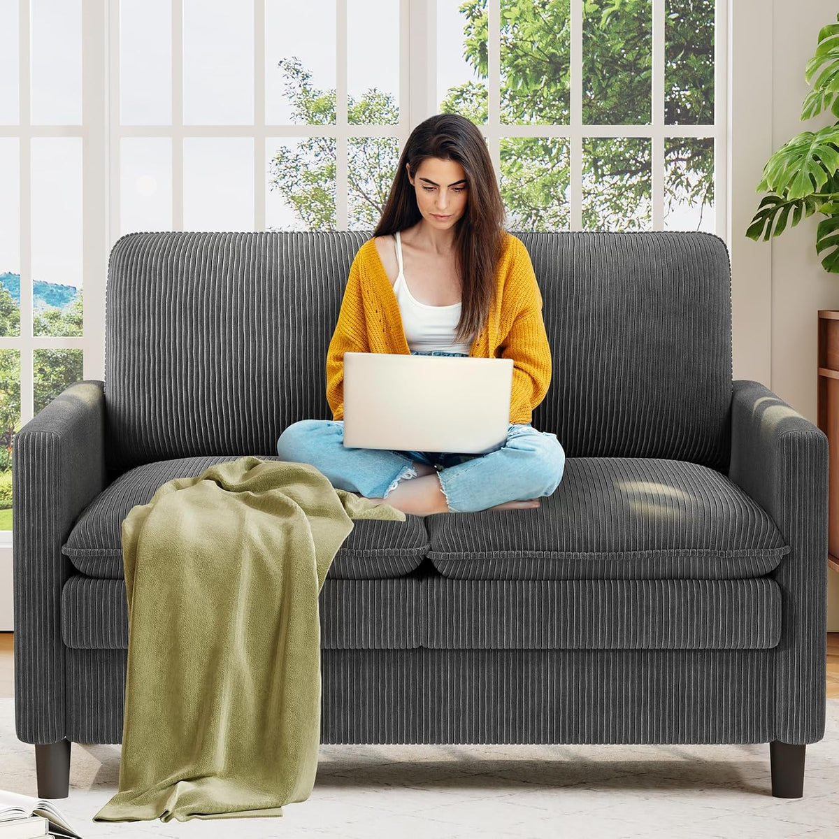 Woman seated cross‑legged with a laptop on a ribbed dark gray loveseat, an olive throw spilling onto the seat.