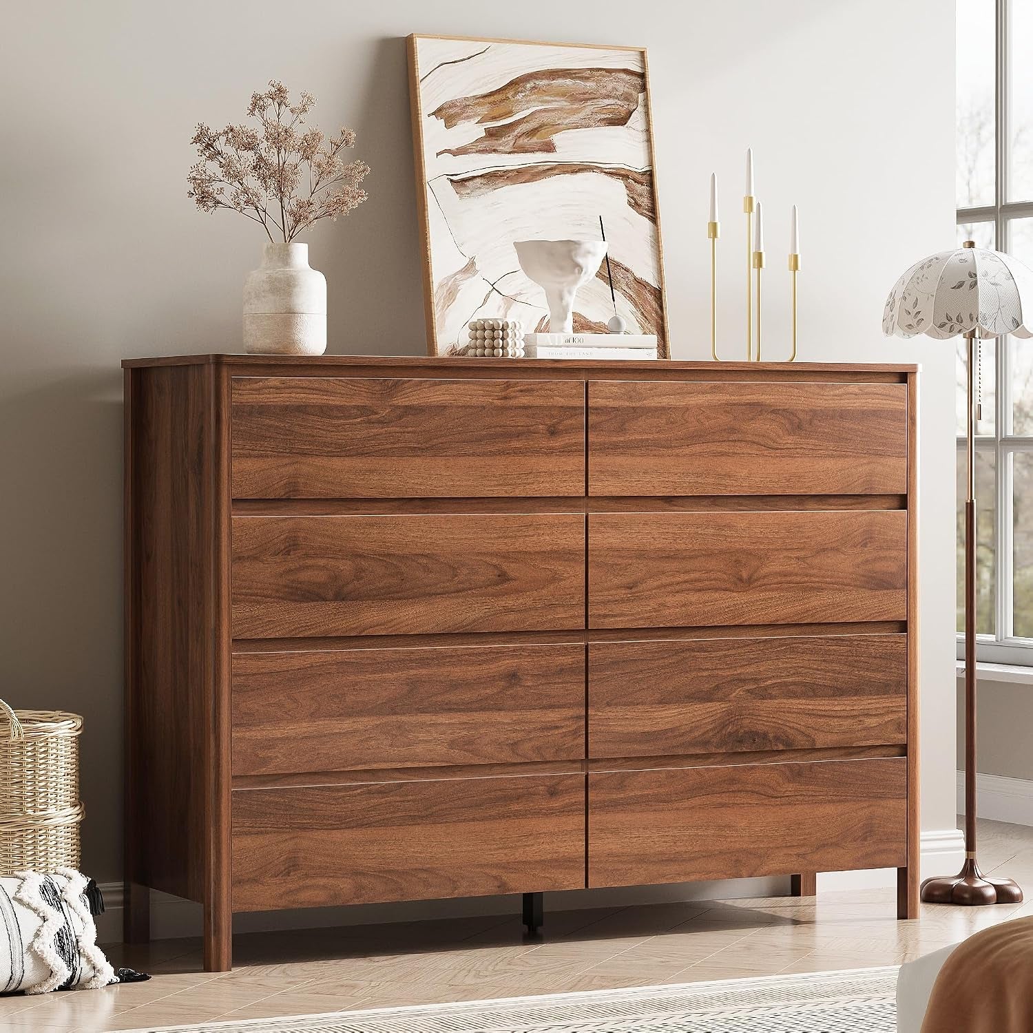 Low-angle shot of a walnut six-drawer dresser with leaning artwork, vase, candlesticks, floor lamp and woven basket.